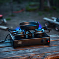 Double burner camp stove with cast iron pan, blue flames, at forest campsite