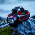 Compact waterproof LED headlamp with red strap accents on a wet mossy rock