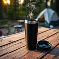 Black insulated stainless steel travel coffee mug with steam, wooden picnic table at sunrise campsite